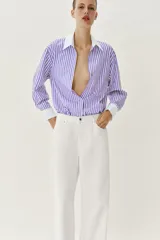 Camisa de popelín con diseño de rayas verticales en tonos blanco y lila, cuello solapa con escote en pico, manga larga y puños anchos contrastantes en color blanco. Cierre frontal con botones metálicos dorados.