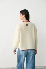 Sweater de tejido bouclé color crudo con detalles de ribete en color rojo en el cuello, puños y bolsillos. Presenta un diseño de manga larga, cuello en V y bolsillos frontales.