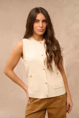 Chaleco de tejido de punto color crema, sin mangas, con cuello redondo y cierre frontal de botones dorados. Cuenta con dos bolsillos frontales aplicados con detalles de botones dorados.