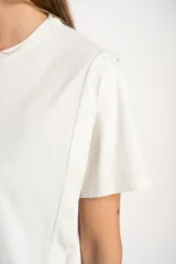 Remera de algodón color mostaza/naranja quemado, con cuello redondo y mangas cortas. Presenta un diseño con costuras externas visibles en los hombros y laterales, dándole un aspecto desestructurado.