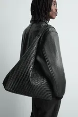 Bolso de hombro tipo hobo o shopper, color negro, con cuerpo de material trenzado. Posee un asa de hombro y un bolsillo interior con cierre de cremallera.