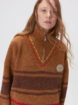 Pullover de punto con textura aterciopelada, diseño oversize, cuello polo con cierre frontal parcial y detalles de pespuntes en contraste. Presenta rayas horizontales en tonos tierra y un parche bordado con la letra M en el pecho.