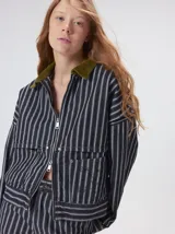 Campera de denim a rayas verticales con cuello de pana en color bordó. Presenta cierre frontal, mangas desmontables con cierre en los hombros, canesú en el pecho y bolsillos de parche cuadrados.