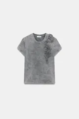 Camiseta de algodón con efecto lavado, cuello redondo y manga corta. Presenta un detalle decorativo de flores en relieve confeccionadas en el mismo tejido sobre el lado izquierdo del pecho.