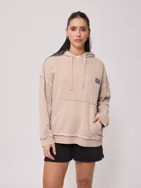 Canguro Rusty con capucha y efecto lavado color beige, de corte oversize, con bolsillo tipo canguro frontal, cordones ajustables y parche de la marca en el pecho. Presenta costuras expuestas y detalles deshilachados.