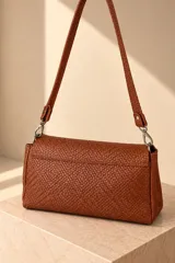 Cartera de cuero vacuno color marrón oscuro, con diseño trenzado y solapa frontal con placa metálica dorada grabada con la marca PECARÍ. Cuenta con correa larga para hombro y herrajes metálicos plateados.