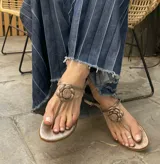 Sandalia plana de cuero color platino metalizado, estilo ojota con tira central y aplique decorativo de flor tridimensional en el empeine. Posee tira fina ajustable en el tobillo.