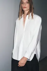 Camisa blanca de popelín de algodón 100%, con cuello solapa, manga larga terminada en puño y cierre frontal con botones ocultos por solapa. Presenta un corte holgado y ruedo curvo.