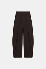 Pantalón de tiro alto color marrón oscuro, con diseño de pierna ancha tipo "balloon" y pliegues en el delantero. Incluye bolsillos laterales ocultos y cierre frontal con cremallera, botón interior y ganchos metálicos.