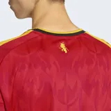 Camiseta de fútbol Adidas de la primera equipación de Bélgica, color rojo con patrón sutil de llamas y demonios. Presenta cuello redondo con ribete negro y amarillo, y detalles de las tres franjas amarillas en los hombros. El escudo de la RBFA está bordado en el pecho.