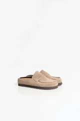 Zuecos tipo mule de gamuza con diseño de mocasín, punta redondeada y tira frontal estilo penny. Presentan costuras decorativas visibles y suela plana de goma.