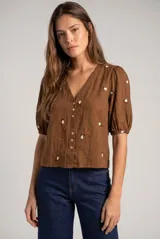 Blusa de viscosa color marrón, con escote en V, mangas cortas abullonadas y cierre frontal con botones. Cuenta con pequeños detalles bordados distribuidos por la prenda.