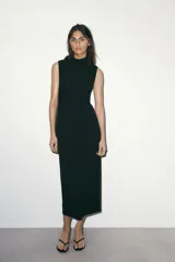Vestido midi de color negro, con cuello subido y diseño sin mangas. Presenta un detalle de drapeado lateral en la parte delantera y cierre de cremallera oculta en la espalda.