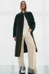 Trench oversize de color verde oscuro, con cuello de piel en tono marrón a contraste. Presenta manga larga con trabillas en los puños, bolsillos delanteros, cierre frontal con cremallera y botones ocultos bajo solapa, y abertura en la espalda.