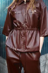 Chaqueta de efecto piel en color bordó, con cuello solapa, cierre frontal con cremallera y mangas por debajo del codo. Presenta cintura elástica y falsos bolsillos delanteros con botones metálicos.