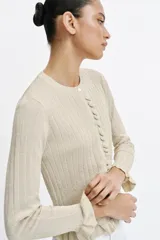Chaqueta de punto canalé en color beige con hilo metalizado, cuello redondo, manga larga y cierre frontal con botones. Presenta detalles de volantes en la tapeta de los botones, el ruedo y los puños.