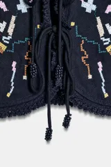 Top corto de color negro con escote en pico y mangas cortas anchas. Presenta bordados coloridos en estilo geométrico y floral, detalle de entredoses y cierre frontal mediante lazos con abalorios.