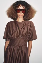 Vestido midi color marrón con textura brillante y arrugada, cuello redondo y mangas cortas tipo murciélago. Presenta un canesú fruncido con pliegues verticales en la cintura que da paso a una falda de corte A.