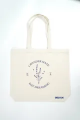 Bolso tipo tote bag de color crudo con estampado de flores celestes y centros azul oscuro. Cuenta con doble asa de hombro en tono crudo y etiqueta con la marca en el frente.