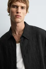 Camisa negra de manga corta con cuello solapa y textura arrugada o plisada. Corte relajado (relaxed fit) con aberturas laterales y cierre frontal de botones.