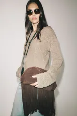 Bolso de mano tipo clutch confeccionado en serraje color marrón, con diseño estructurado y cierre a presión. Presenta un detalle de flecos largos en la parte inferior y cuenta con forro y bolsillo interior.