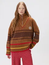 Pullover de punto con textura aterciopelada, diseño oversize, cuello polo con cierre frontal parcial y detalles de pespuntes en contraste. Presenta rayas horizontales en tonos tierra y un parche bordado con la letra M en el pecho.