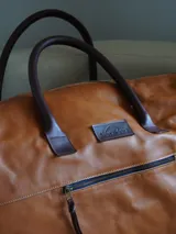 Bolso tipo weekender de cuero marrón, con asas de mano y bolsillo frontal con cierre metálico.