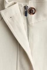 Campera corta de lino color marfil con cuello alto, solapa con botones, bolsillos diagonales con solapa, mangas raglán y cordón ajustable en la cintura.