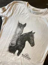 Remera blanca de manga corta con cuello redondo, confeccionada en algodón. Presenta un estampado fotográfico en tono gris oscuro de dos caballos junto al isotipo de la marca en la parte inferior y un aplique marrón en la manga.