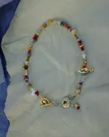 Collar corto de estilo artesanal, compuesto por una variedad de piedras naturales en tonos tierra, verdes, rojos y amarillos, intercaladas con pequeñas perlas de río. Presenta dijes de bronce macizo con formas orgánicas, incluyendo una figura de pez y un dije circular central, unidos por un cierre de barra y argolla.