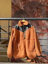 Canguro de peluche color marrón anaranjado con cuello alto y detalles en cuero sintético negro en la parte superior del pecho y cuello. Tiene cierre parcial con botones a presión y dos bolsillos frontales.