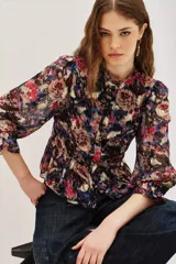 Camisa de manga larga con estampado floral y detalles en hilo metalizado. Presenta cuello alto con volados, cierre frontal con botones y puños con volados. El diseño incluye un corte entallado en la cintura con un ligero peplum.