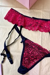 Conjunto de lencería compuesto por una tanga y un portaligas, confeccionados en encaje color fucsia y detalles en negro. La tanga presenta un diseño de encaje frontal con terminaciones en lycra negra y un pequeño lazo decorativo. El portaligas cuenta con una banda ancha de encaje y tiras ajustables con ganchos metálicos.