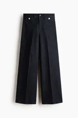 Jean tobillero de denim azul oscuro, de tiro alto y piernas amplias con corte holgado. Presenta bolsillos delanteros tipo parche con solapa y botones dorados, además de pliegues marcados en el frente.