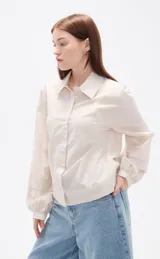 Camisa de algodón con estampado de rayas verticales finas en tonos beige y blanco, cuello clásico, cierre frontal con botones y mangas largas con puños anchos estructurados.