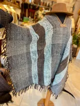 Poncho de lana virgen tejido en telar con diseño geométrico a rayas en tonos gris, marrón y celeste, con apliques de cuero en el escote y flecos en el ruedo.