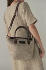 Cartera tipo bolso de mano confeccionada en lana fina sin teñir, con detalles en cuero marrón oscuro en las asas y correa ajustable. Incluye cierre superior y etiqueta de cuero con logo.