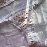 Conjunto de lencería de dos piezas confeccionado en tela acanalada blanca con estampado de pequeñas flores rosas. Incluye un soutien tipo triángulo sin aro, con detalles de encaje blanco en los bordes y una argolla metálica en el centro, y una bombacha colaless ajustable con tiras laterales y detalles de encaje.