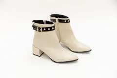 Bota corta color beige de cuero, con punta fina y taco cuadrado. Presenta aplique de charol negro con hebilla dorada y tachas pequeñas alrededor del tobillo. Cuenta con cierre lateral.