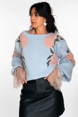 Suéter de punto color celeste con estampado floral en tonos rosados y verdes. Presenta mangas amplias con puños de encaje en color nude y cuello redondo.