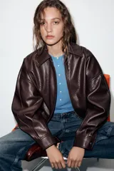 Cazadora estilo bomber de efecto piel en color bordó, con cuello solapa, manga larga, bolsillos delanteros de vivo con botón a presión y bajo elástico. Cierre frontal con cremallera oculta y botones a presión.