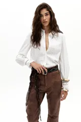 Camisa blanca de popelina de algodón con alforzas en el frente, cuello con terminación ondulada y bordados, y puños con vainillas. Tiene un corte levemente entallado, ruedo curvo y un sutil frunce en los hombros.