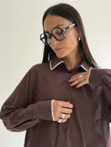 Camisa oversize de color marrón con cuello camisero y puños con detalle de bordado festoneado en color blanco. Presenta cierre frontal de botones y mangas largas.