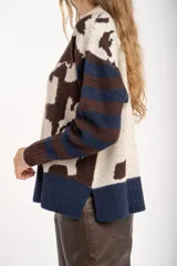 Sweater de punto con diseño combinado, presentando un estampado de manchas de vaca en el cuerpo y mangas con rayas horizontales en tonos azul y marrón. Posee cuello redondo y puños acanalados.