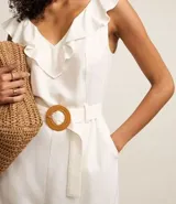 Enterito corto blanco sin mangas confeccionado en viscolino con escote V, espalda descotada con volados en el escote. Posee cinturón con hebilla redonda de ráfia y trabillas en la cintura.