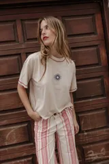 Remera de color beige con cuello redondo y manga corta, que presenta un bordado central en forma de sol y costuras decorativas a contraste en color rojo en el cuello, mangas y laterales.