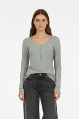 Remera blanca de manga larga, entallada, con cuello redondo y una hilera de botones en el frente. Incluye un pequeño bolsillo decorativo en el pecho.