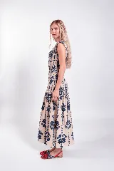 Vestido largo sin mangas de color beige con estampado floral y vegetal en color azul marino. Cuenta con un cuerpo ajustado mediante fruncidos elásticos (smock) y falda larga con vuelo y corte en capas.