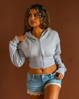 Campera tipo hoodie corta con cierre frontal, confeccionada en tela de algodón color celeste. Presenta un bordado en hilo rosado en el pecho y cintura ajustada con elástico acanalado.