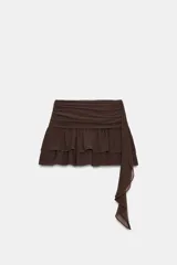 Falda pantalón de tiro bajo en color negro, con cintura drapeada y detalle de tira lateral colgante. Presenta un diseño con volantes en el bajo y cierre de cremallera metálica.